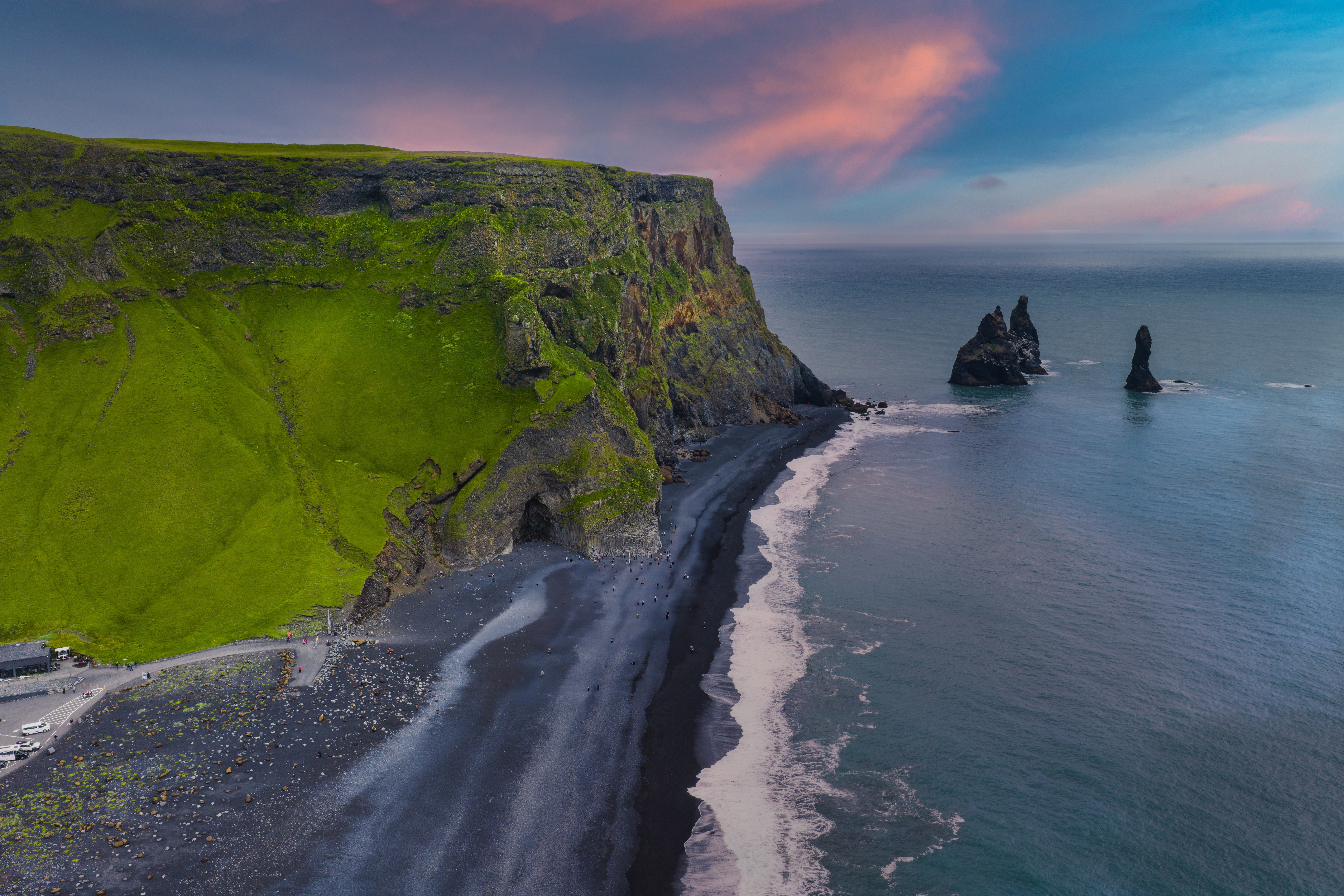 Aerial view of a green clifftop coastline and black-sand beach at sunset